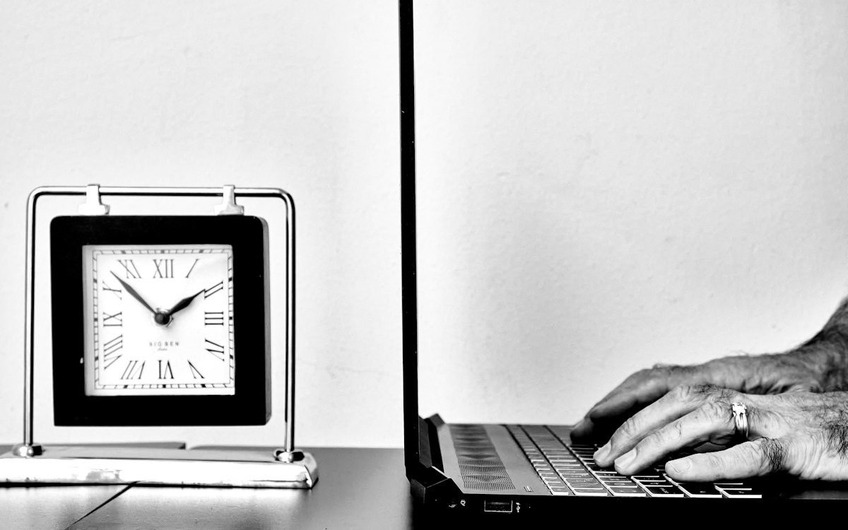 Relógio sobre a mesa ao lado de uma pessoa trabalhando no notebook
